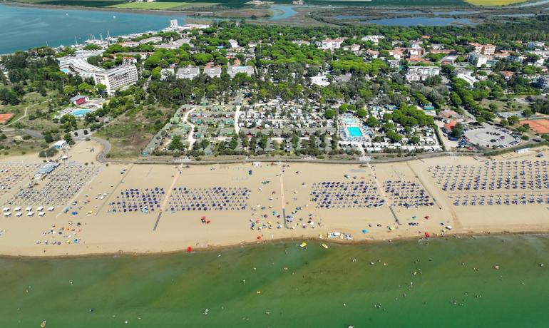 Überfüllter Strand mit Sonnenschirmen und Blick auf baumbestandenen Campingplatz.