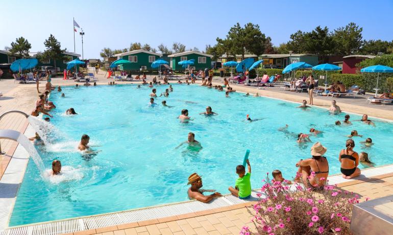 Crowded pool with swimmers, blue umbrellas, and cabins in the background.